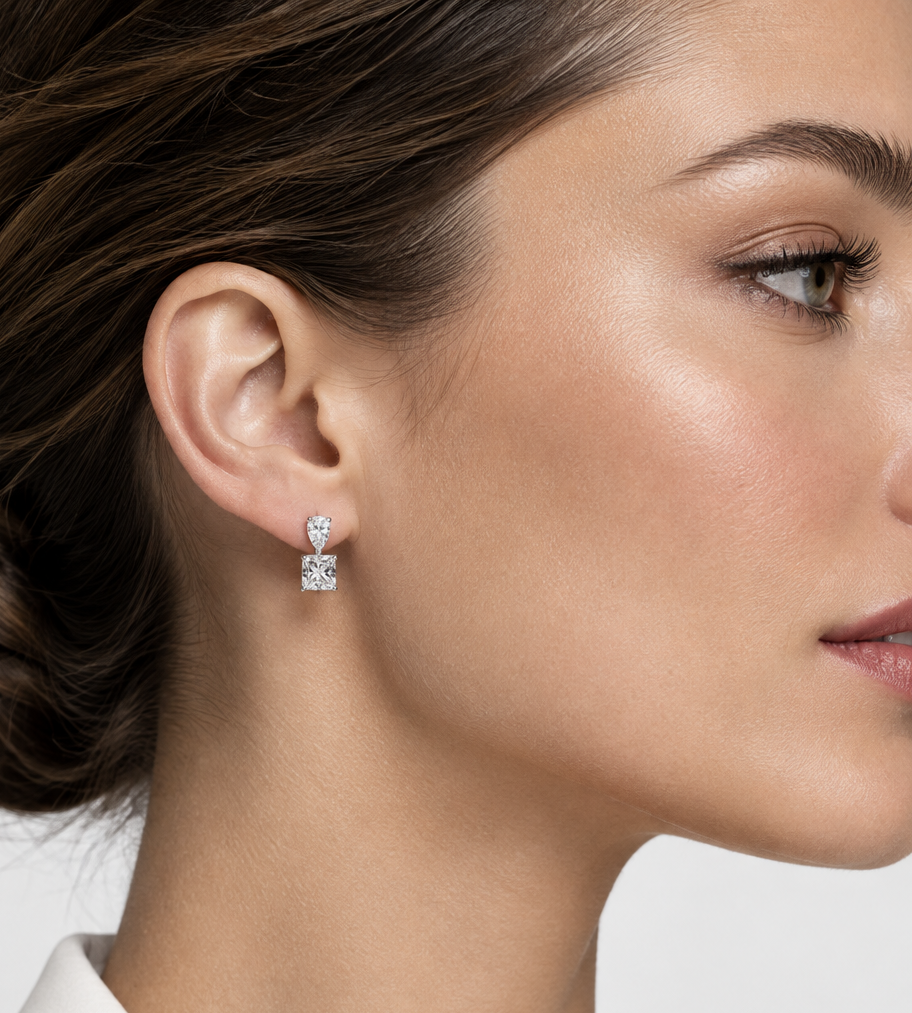 A close-up side profile of a woman wearing a small silver stud earring with a clear square-cut gemstone. The earring features a small silver top and a larger square-cut clear stone below, set against a clean light gray background.